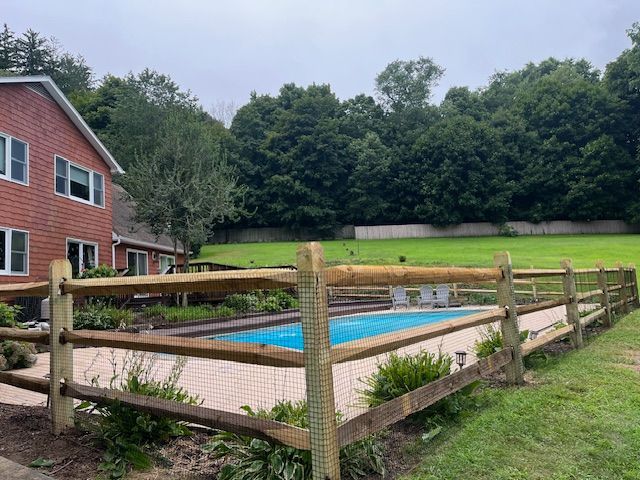 Backyard pool with blue cover beside a red house and wooden fence, surrounded by green lawn and trees