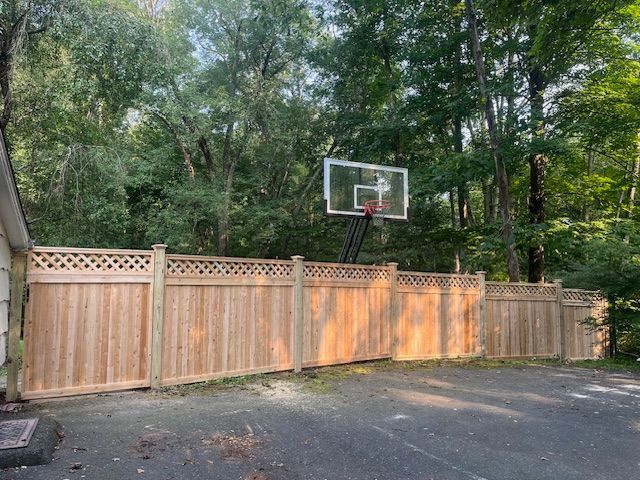 Wooden fence and driveway with a basketball hoop in a wooded backyard.