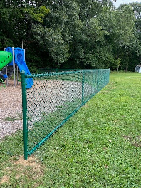 Green chain-link fence beside a playground with a blue slide and grassy park area
