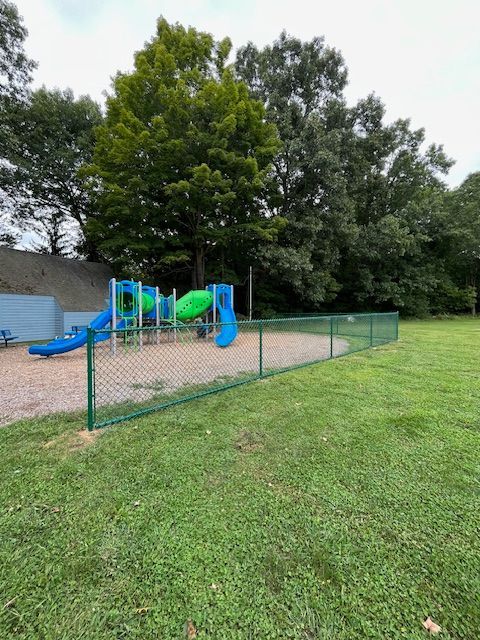 Small playground with blue and green slides and climbing structures beside a grassy field and trees.