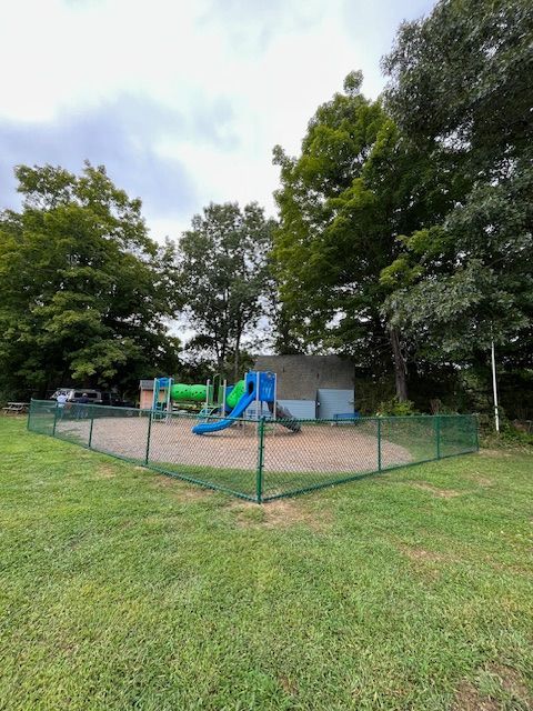 Fenced playground with blue and green play structures on a grassy park lawn, surrounded by trees