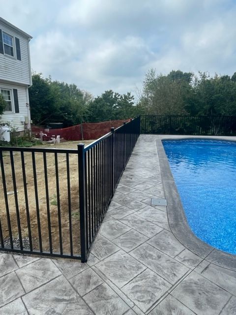 Backyard pool with black fence, stamped concrete deck, and house beside water under cloudy sky