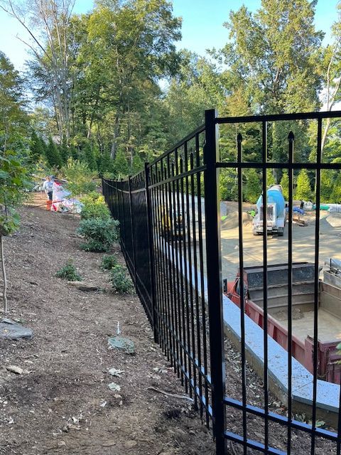 Black metal fence beside a wooded path overlooking a circular concrete structure with blue tarp and workers in the distance