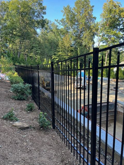 Black metal fence along a sloped dirt path beside a blue truck and trees in a sunny yard