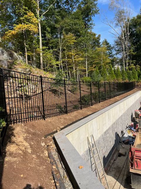 Black metal fence along a wooded hillside beside a concrete drainage channel and construction equipment