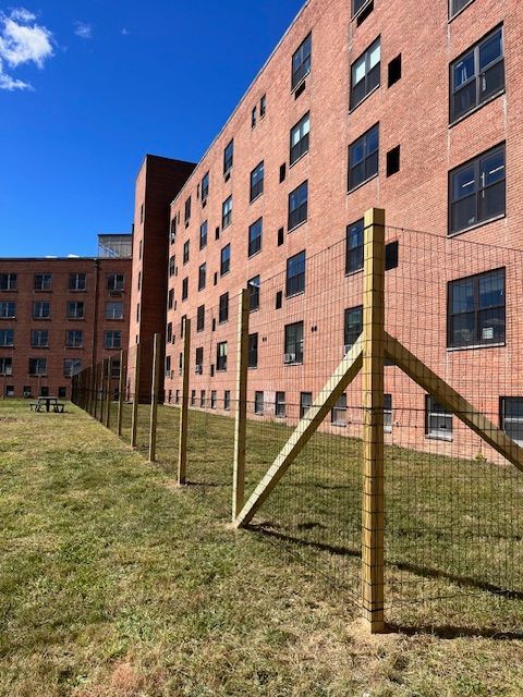 Brick apartment building beside a grassy yard with a wire fence under a blue sky