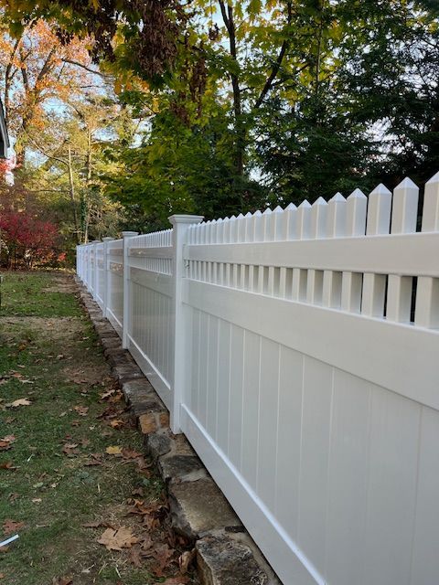 White vinyl picket fence along a leafy yard with autumn trees in the background