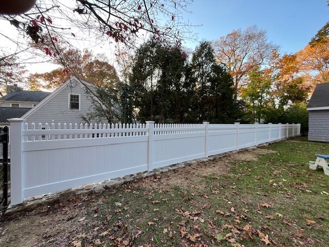 White vinyl fence along a grassy yard beside a house with autumn trees in the background