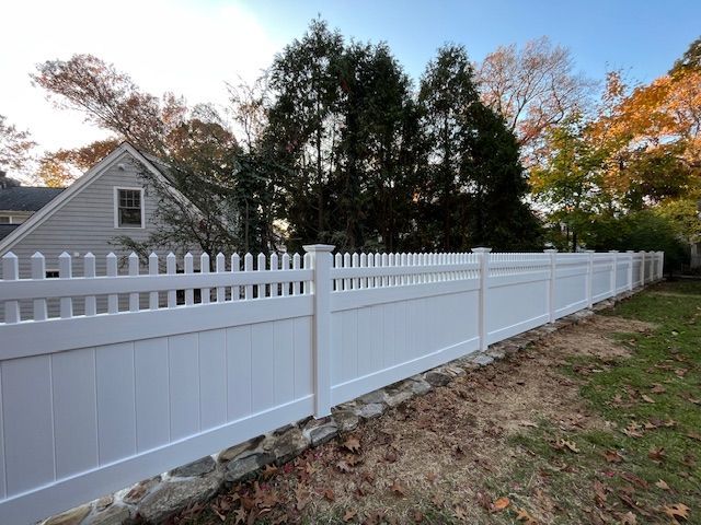 White vinyl picket fence along a yard beside a gray house, with trees and autumn leaves in the background.