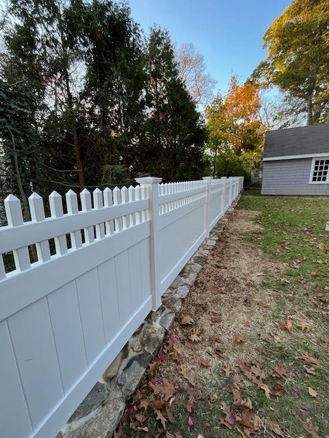 White vinyl picket fence along a yard beside a house, with autumn trees and fallen leaves.