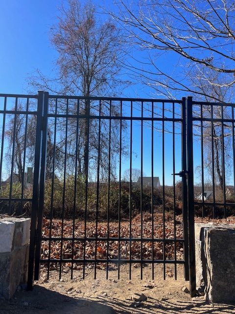 Black metal gate between stone pillars, opening to a wooded path under a clear blue sky.