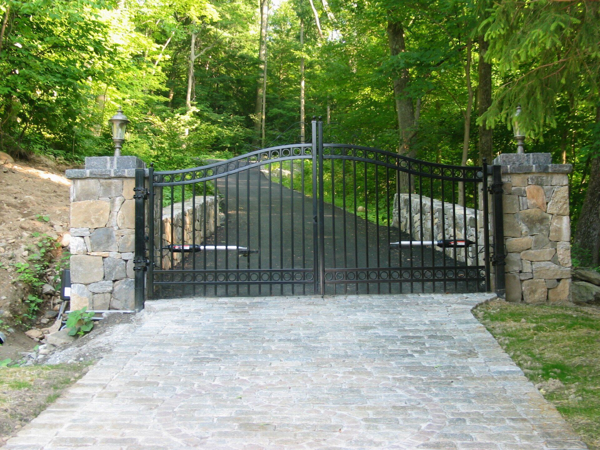 Black iron driveway gate between stone pillars at the end of a paved path in a wooded area