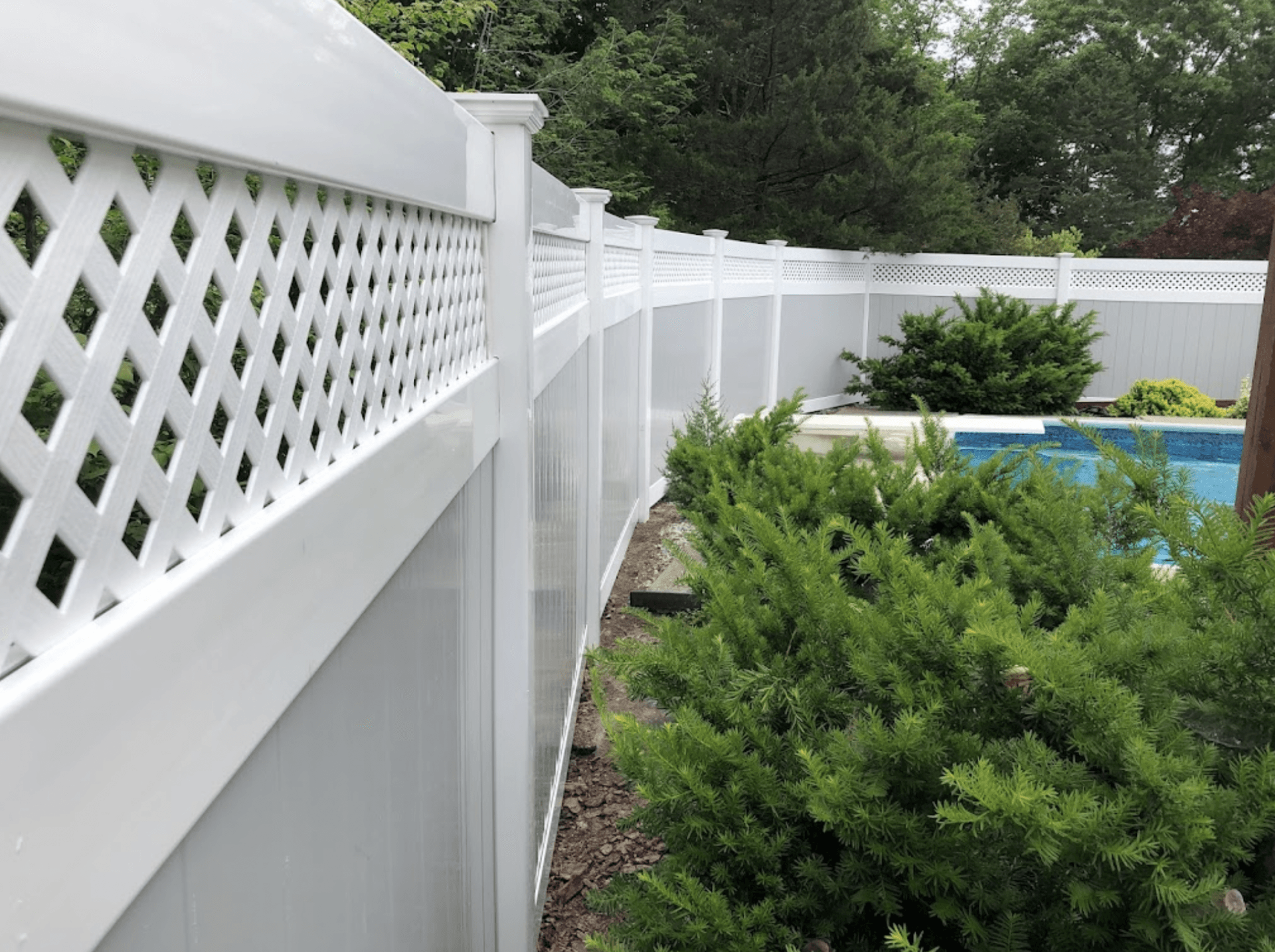 White lattice fence beside a narrow garden path and blue pool, lined with green shrubs and trees.