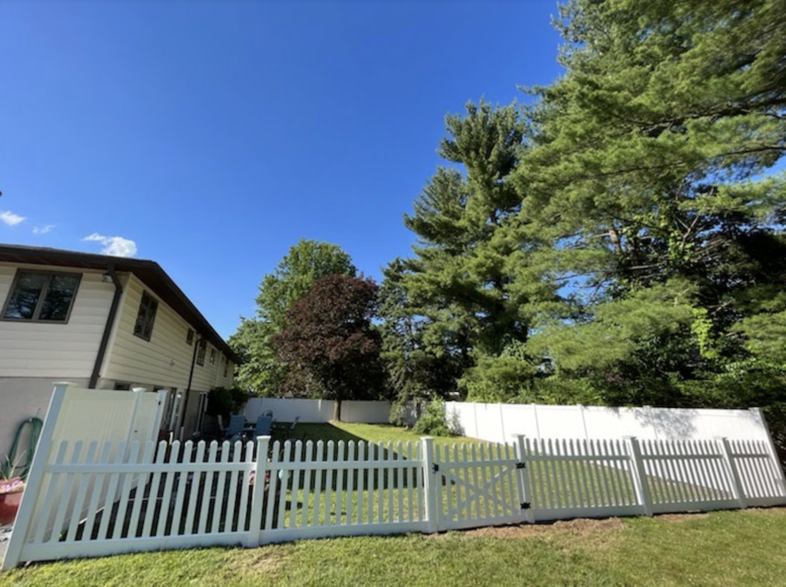 Suburban house with white picket fence, green lawn, and tall trees under a clear blue sky