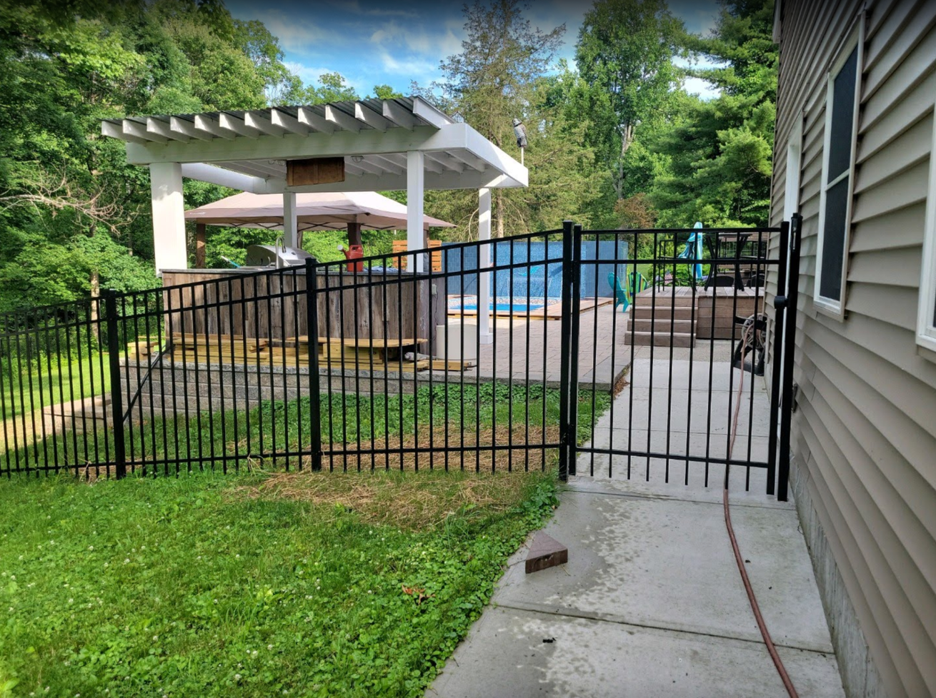 Backyard patio with black metal fence, white pergola, and blue above-ground pool beside a house.