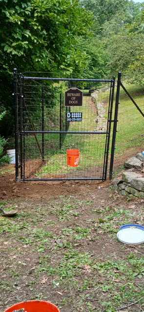 Black metal gate with a warning sign blocks a wooded path; orange bucket hangs below.