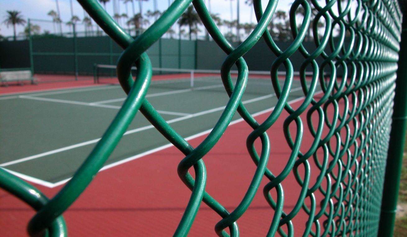 Close-up of a green chain-link fence beside an empty tennis court with red and green surface.