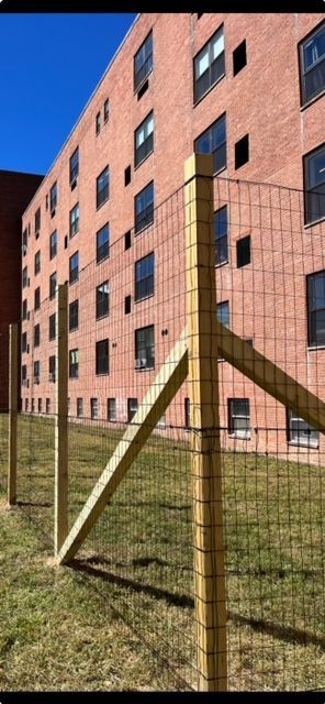 Brick apartment building behind a wire fence and grassy yard under a clear sky