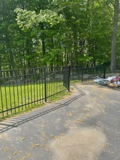 Black metal fence beside a paved path, with trees and trash bags near the wooded edge