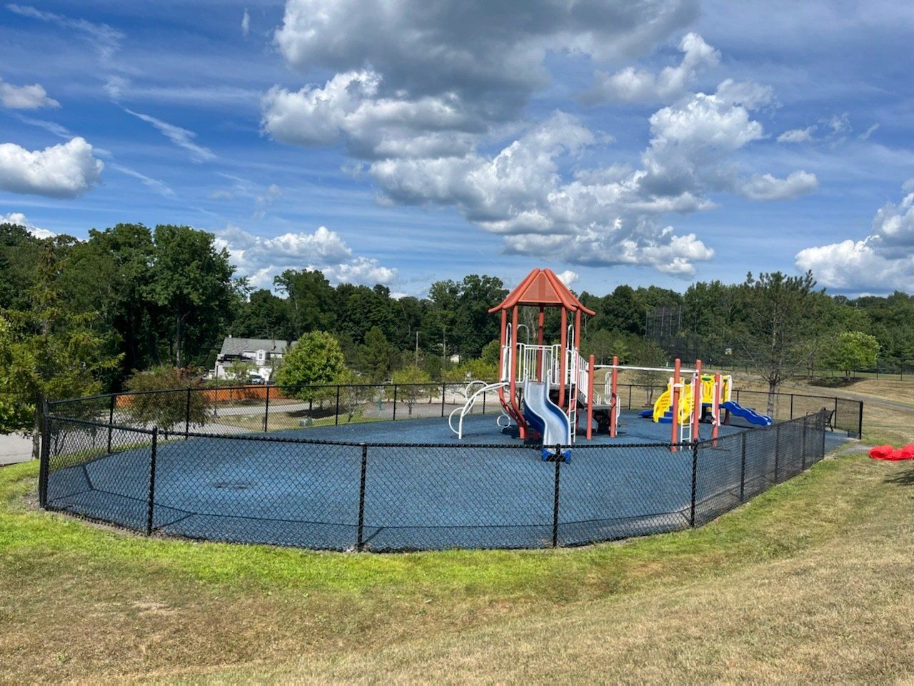 Playground with colorful slides and climbing structure inside a fenced area under a cloudy blue sky