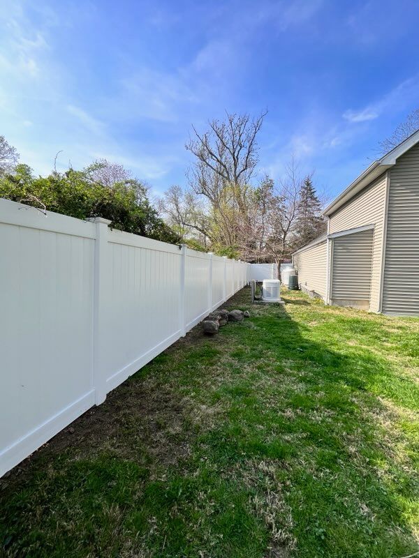 Narrow grassy side yard with a white vinyl fence and beige house under a blue sky