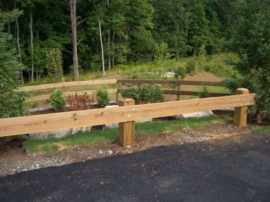 Wooden roadside guardrail beside a landscaped gravel area with trees in the background