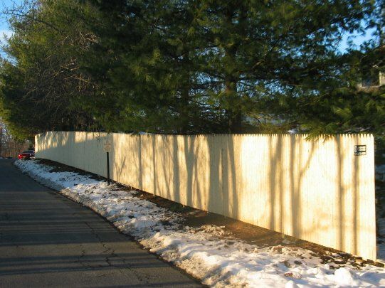 Long beige fence along a road, lined with snow and shaded by tall trees.