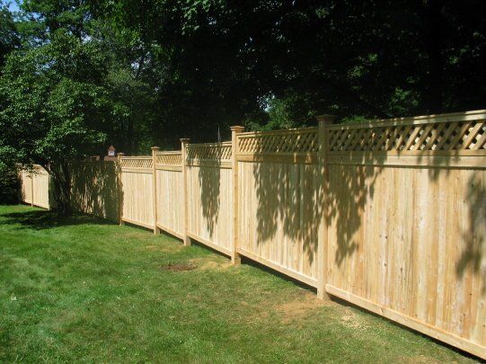 Long wooden fence beside a green lawn under leafy trees, with sunlight casting shadows.