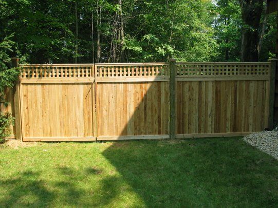 Wooden privacy fence with lattice top in a leafy backyard, bordered by grass and gravel.