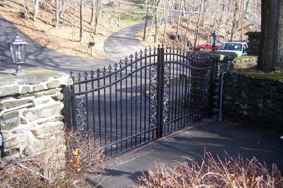 Black metal gate at the end of a stone wall on a paved driveway, with hillside and parked cars beyond