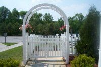 White garden arbor and picket gate on a paved path beside green shrubs and trees under a blue sky