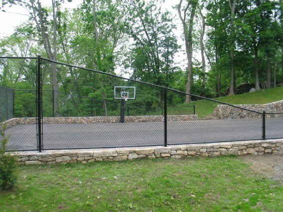 Outdoor basketball court with a single hoop, surrounded by a black fence and trees.