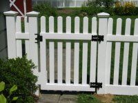 White picket garden gate with black hinges and latch between fence panels, bordered by greenery