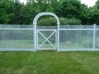 White picket fence with an arched gate and cross-braced center, set in a grassy yard with trees behind it