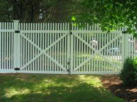 White picket fence with a central gate and diagonal braces in a green, tree-lined yard