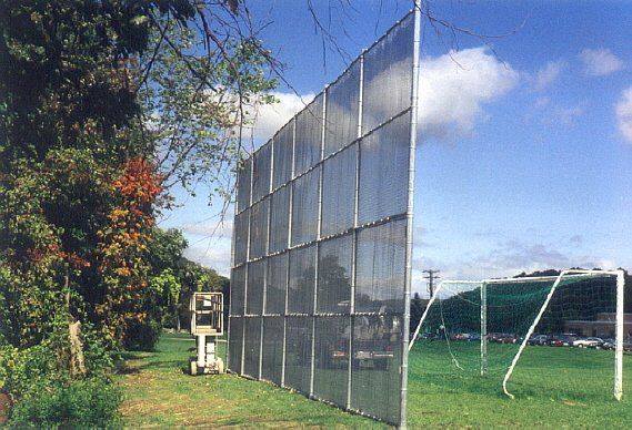 Mesh sports fence beside a grassy field under a blue sky, with trees on the left.