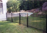 Black metal fence along a grassy yard beside a white house and a rocky wooded slope.