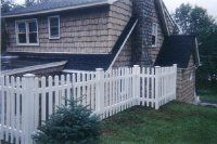White picket fence in front of a shingled house with a small evergreen tree and grassy yard.