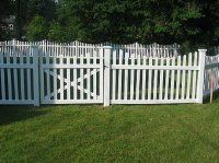 White picket fence with a central gate in a grassy yard, backed by trees and shrubs