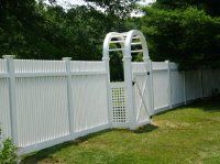 White garden fence with an arched gate in a grassy yard, backed by trees.