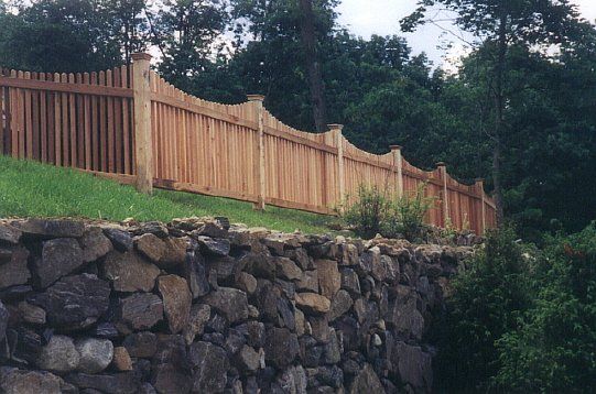 Wooden fence atop a stone retaining wall along a grassy hillside with trees behind it
