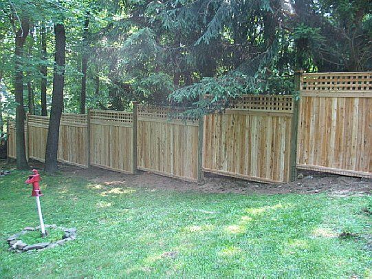 Wooden privacy fence along a grassy yard with tall trees and a red sprinkler in the foreground