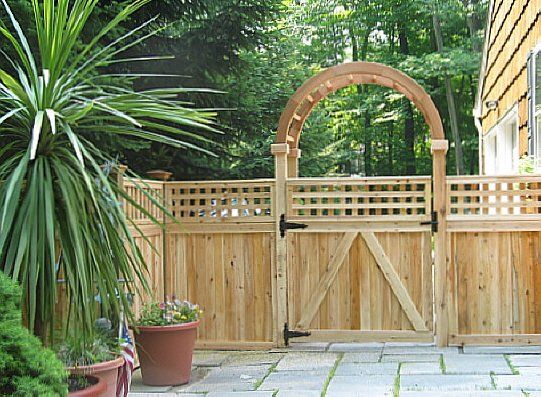 Wooden garden gate with arched trellis, fence, potted plant, and stone patio beside a house