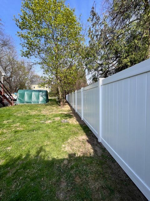 Backyard with grass, trees, and a white vinyl fence under a sunny sky.