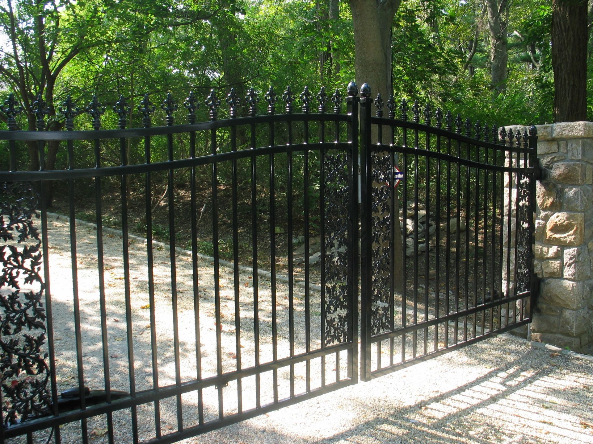 Black metal driveway gate beside a stone pillar, opening onto a wooded path