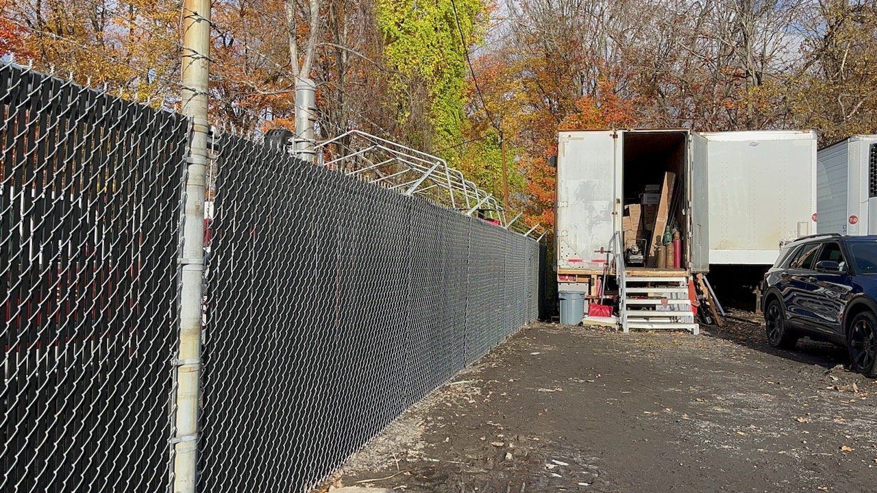 Outdoor lot with chain-link fence, open white truck trailer, and a parked black car in autumn trees