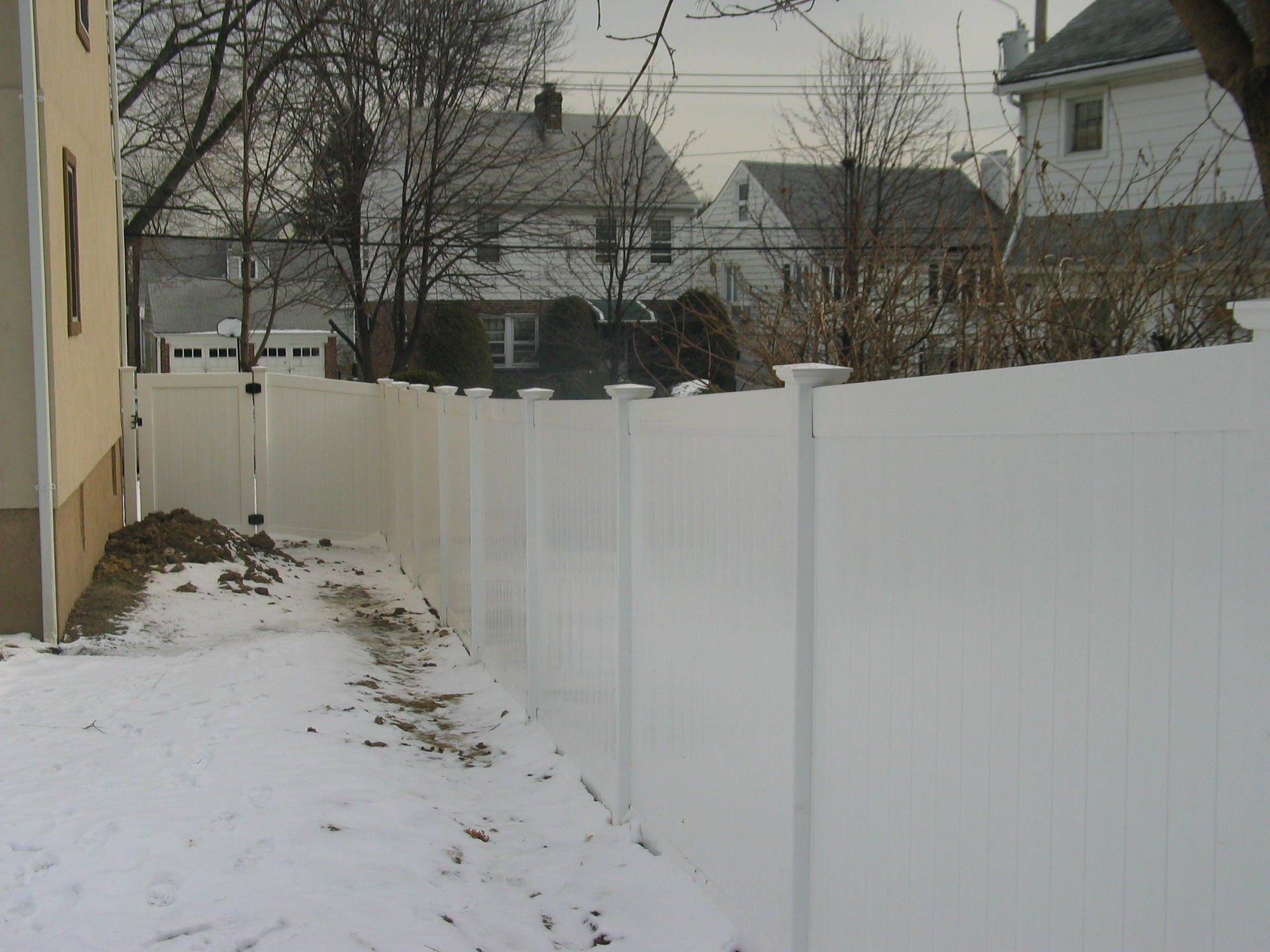 Snowy side yard with a long white fence and houses in the background