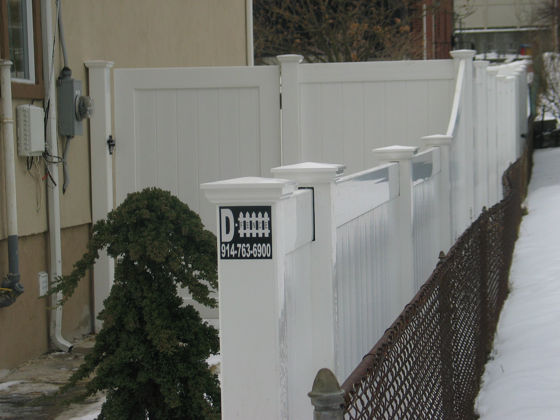 Snowy narrow walkway beside a white fence and house, with a small evergreen shrub and a lattice gate.