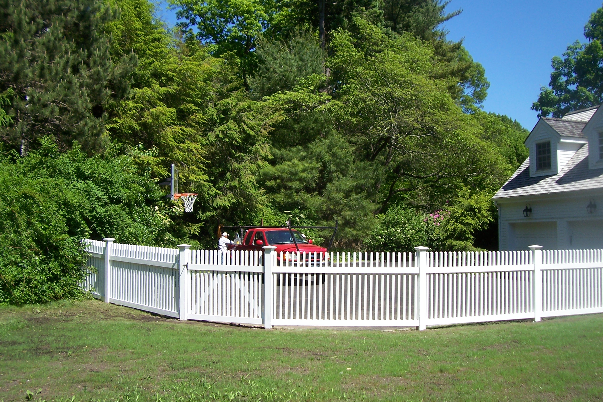 White picket fence around a green yard with a red car and house beside trees