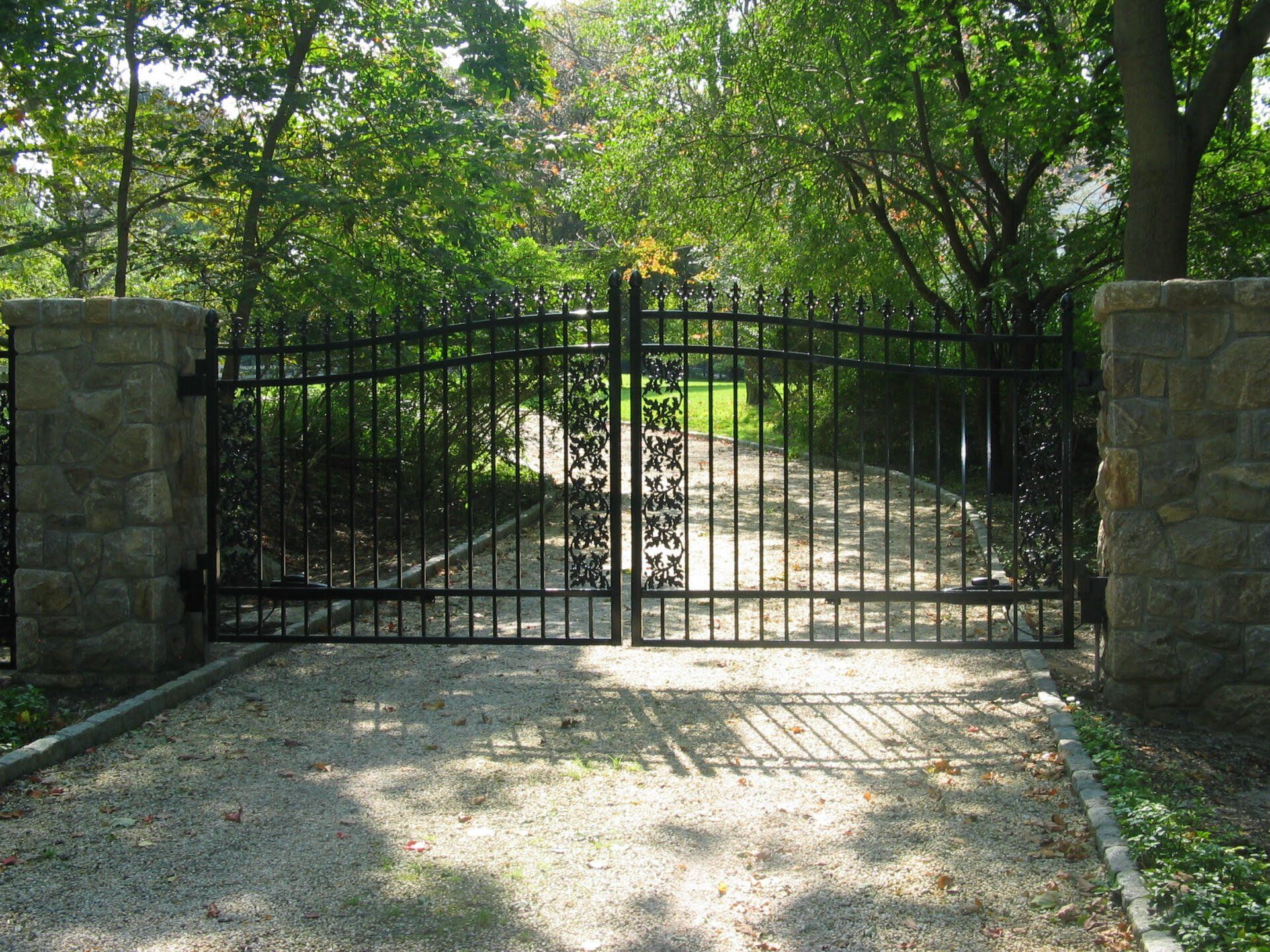 Black metal gate between stone pillars across a sunlit driveway, with trees and greenery behind it